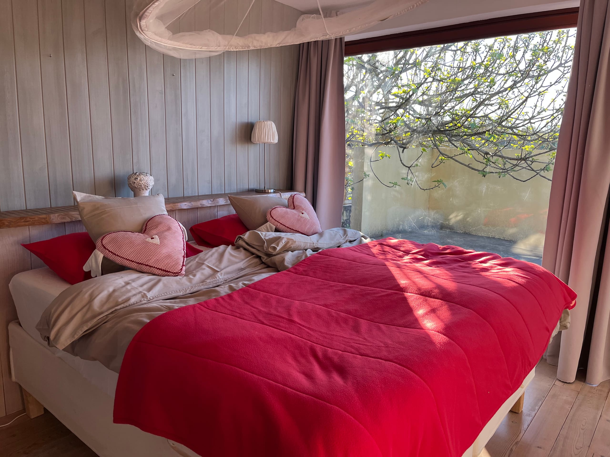 Schlafzimmer mit rotem Bettbezug, herzförmigen Kissen und Moskitonetz vor großem Fenster.