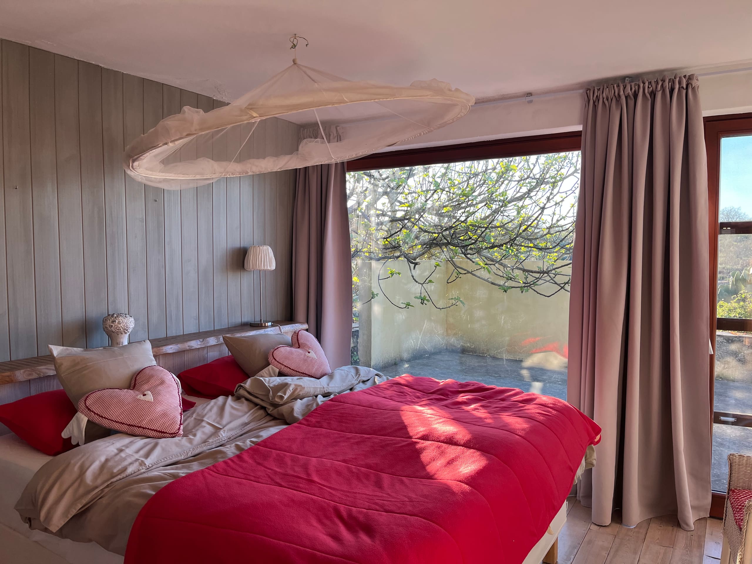 Schlafzimmer mit rotem Bettbezug, herzförmigen Kissen und großem Fenster zur Terrasse.