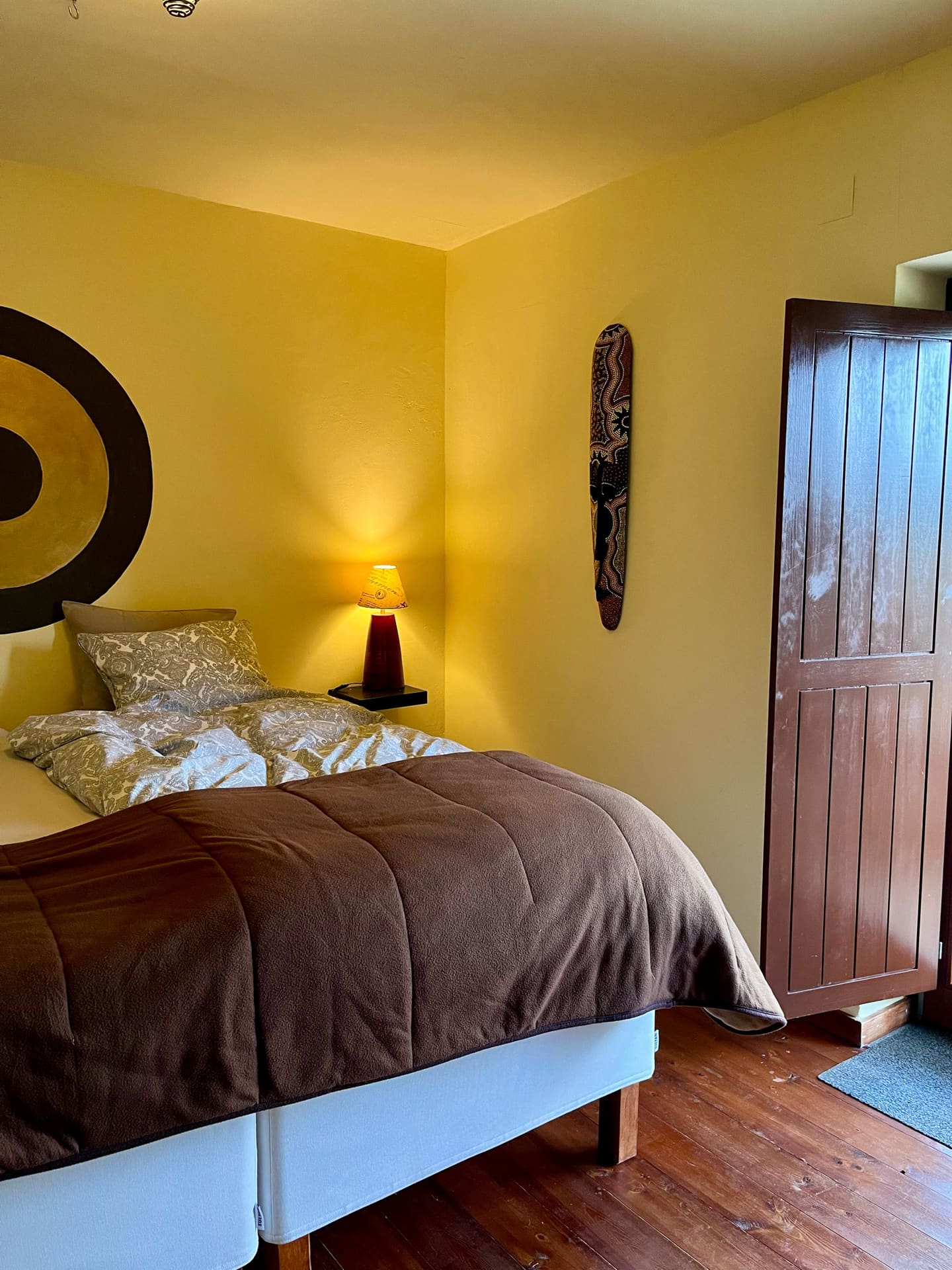 Schlafzimmer mit gelben Wänden, brauner Tagesdecke und offenem Holzschrank.
