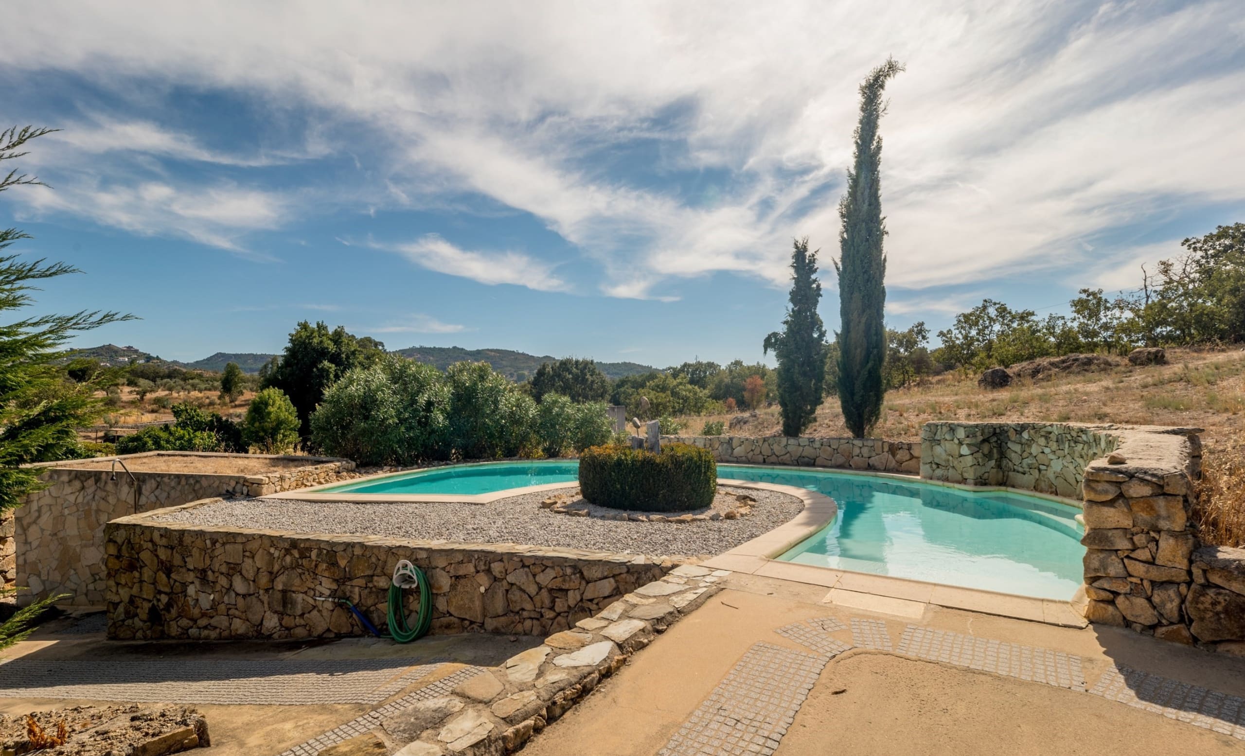 Pool mit Steinmauern und Zypressen vor hügeliger Landschaft