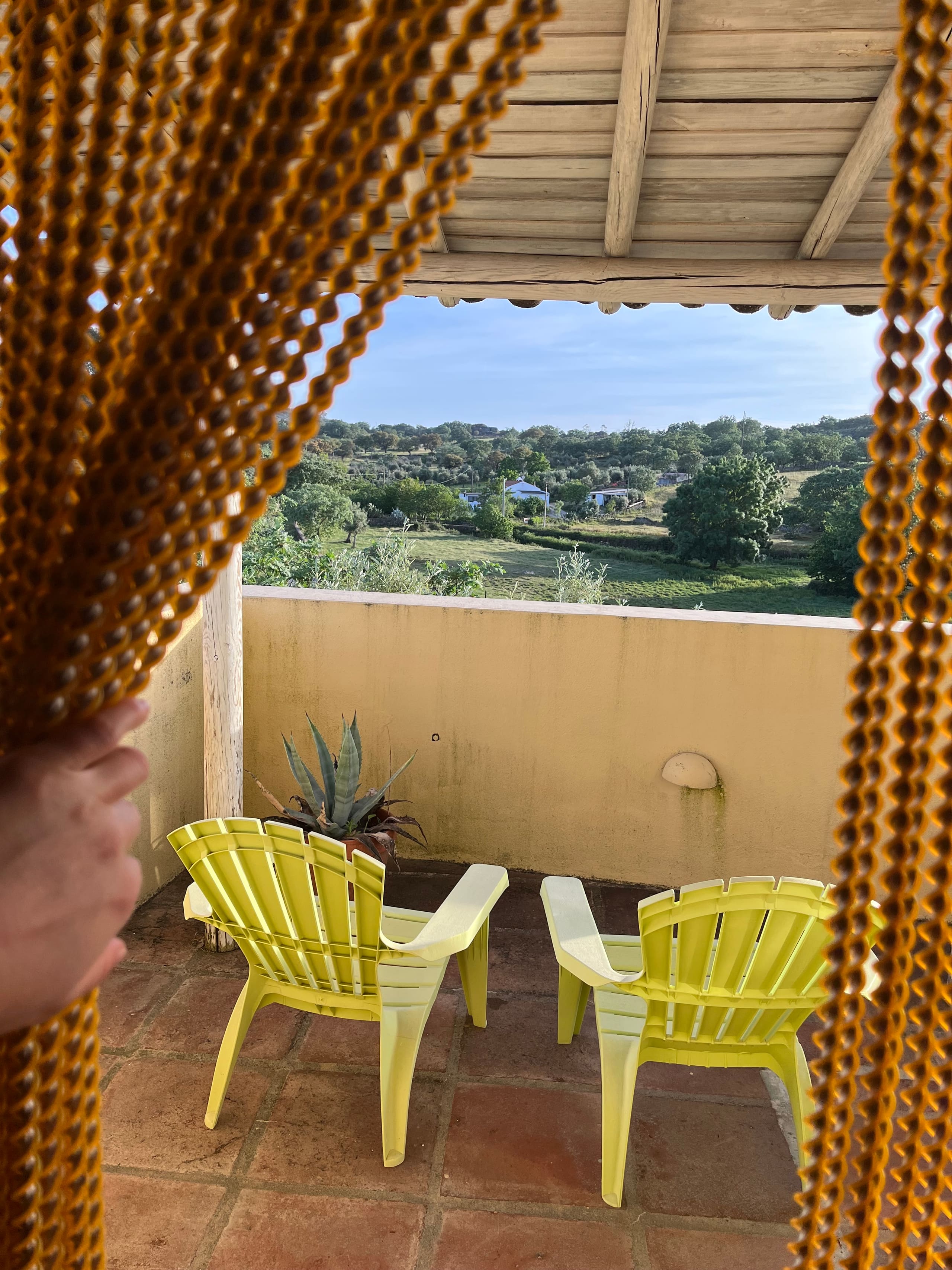 Blick von überdachter Terrasse mit gelben Stühlen über grüne Hügellandschaft.