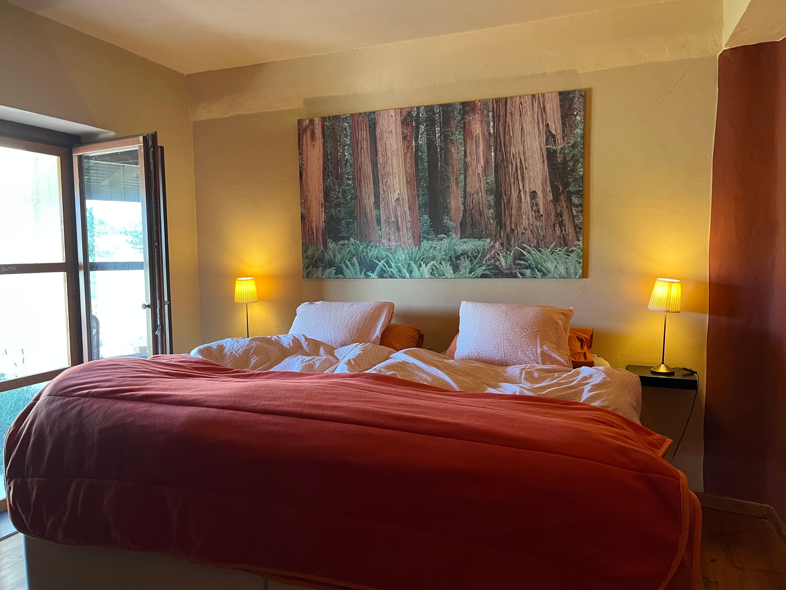 Schlafzimmer mit Doppelbett in Terrakotta-Rot, Nachttischlampen und Waldmotiv über dem Bett.