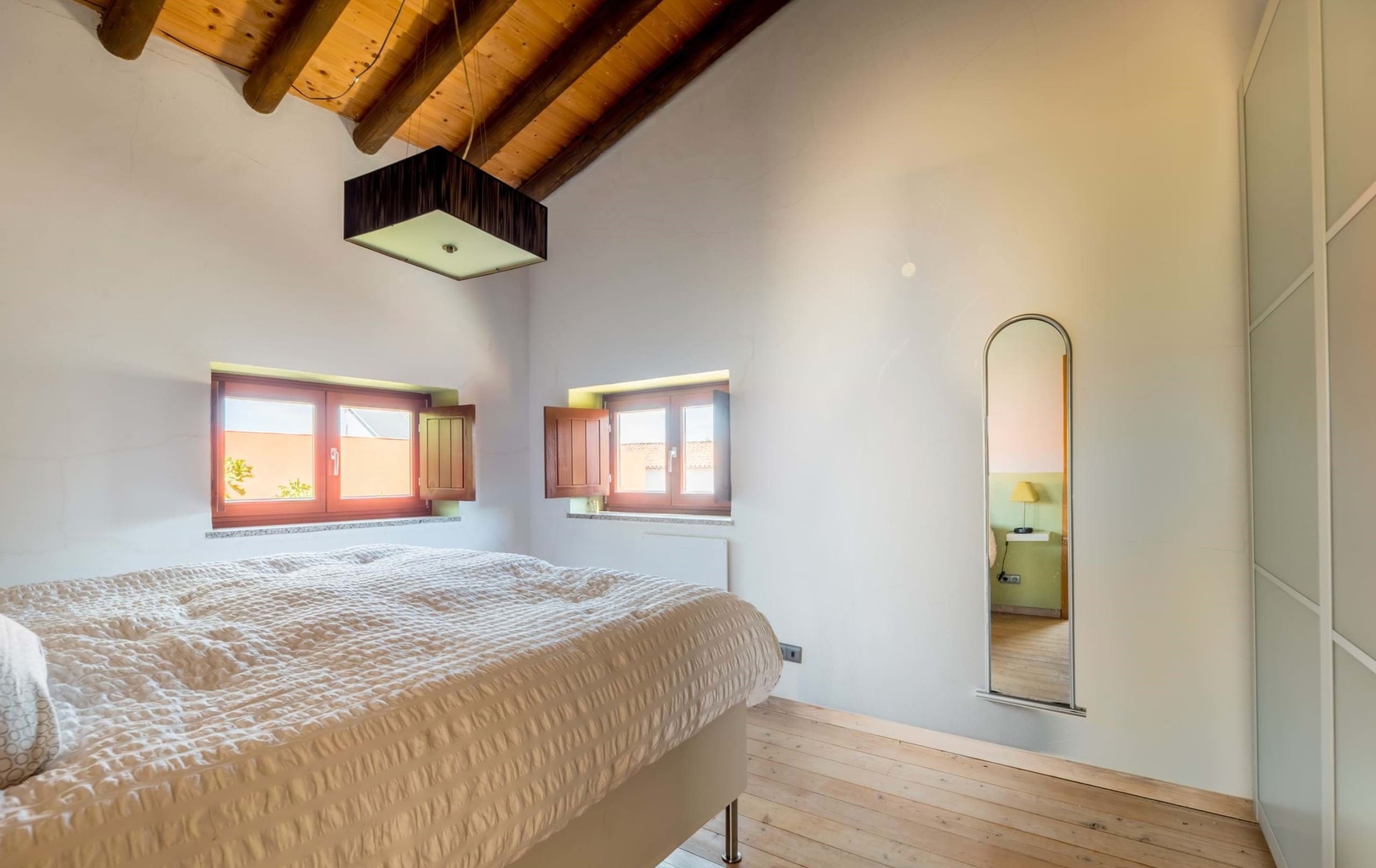 Schlafzimmer mit Holzbalkendecke, Doppelbett, kleinen Fenstern und Wandspiegel.