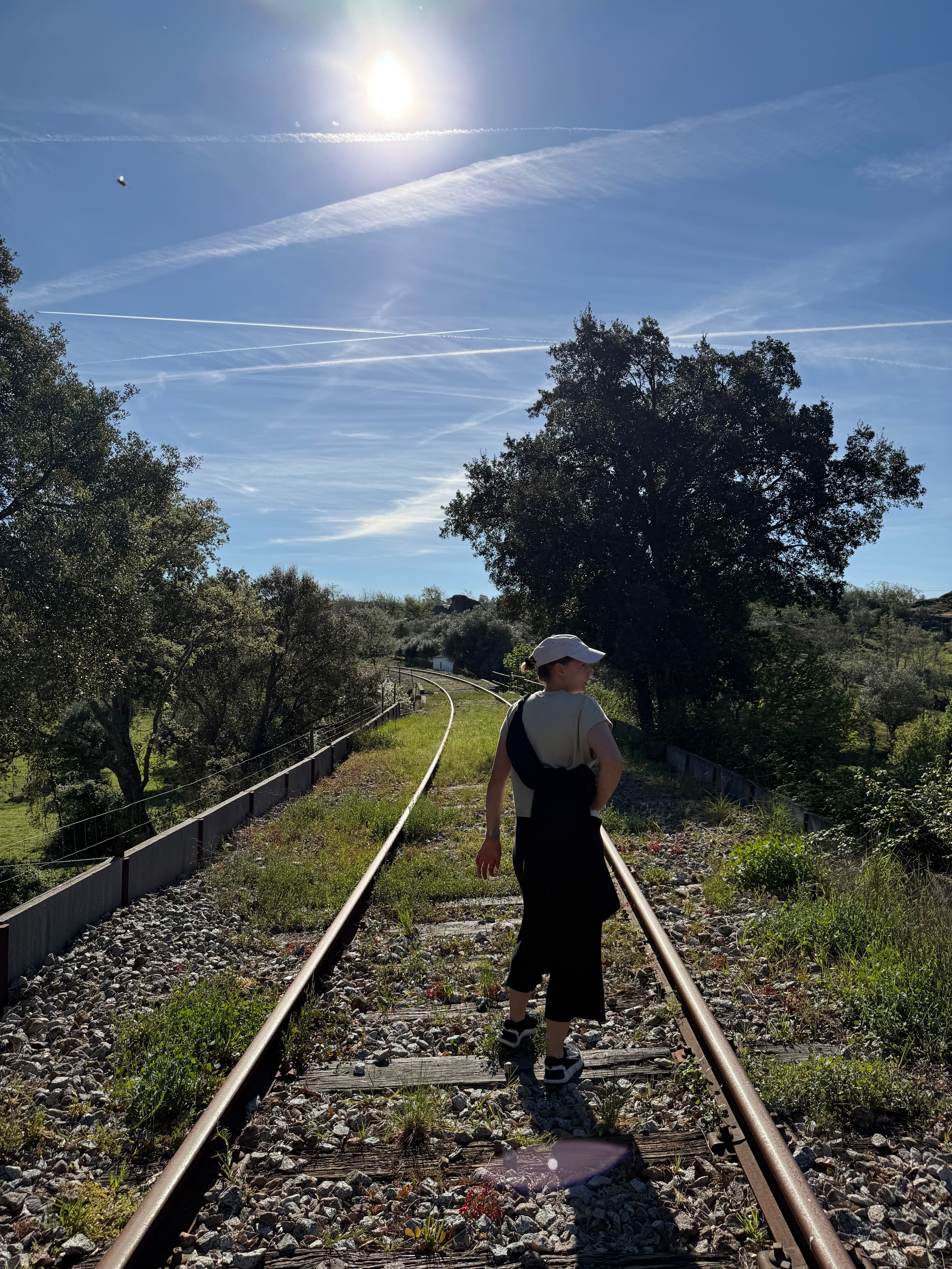 Person geht über stillgelegte Bahngleise durch sonnige, baumgesäumte Landschaft.