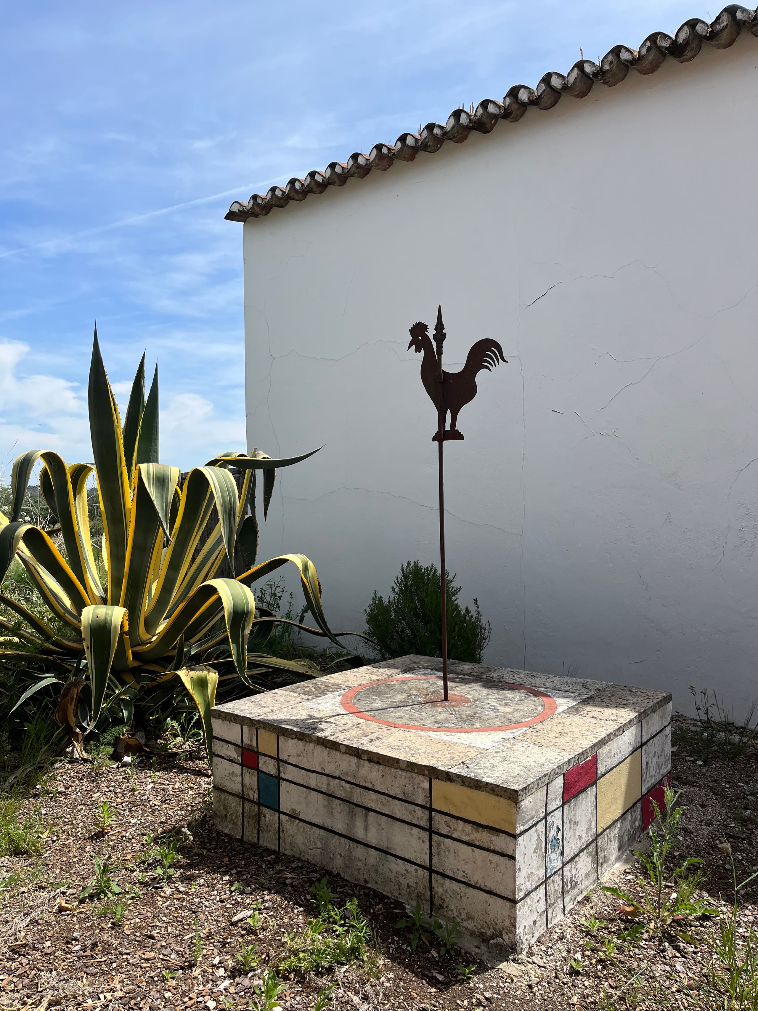 Rostige Hahn-Wetterfahne auf bunt bemaltem Steinsockel neben einer Agave.